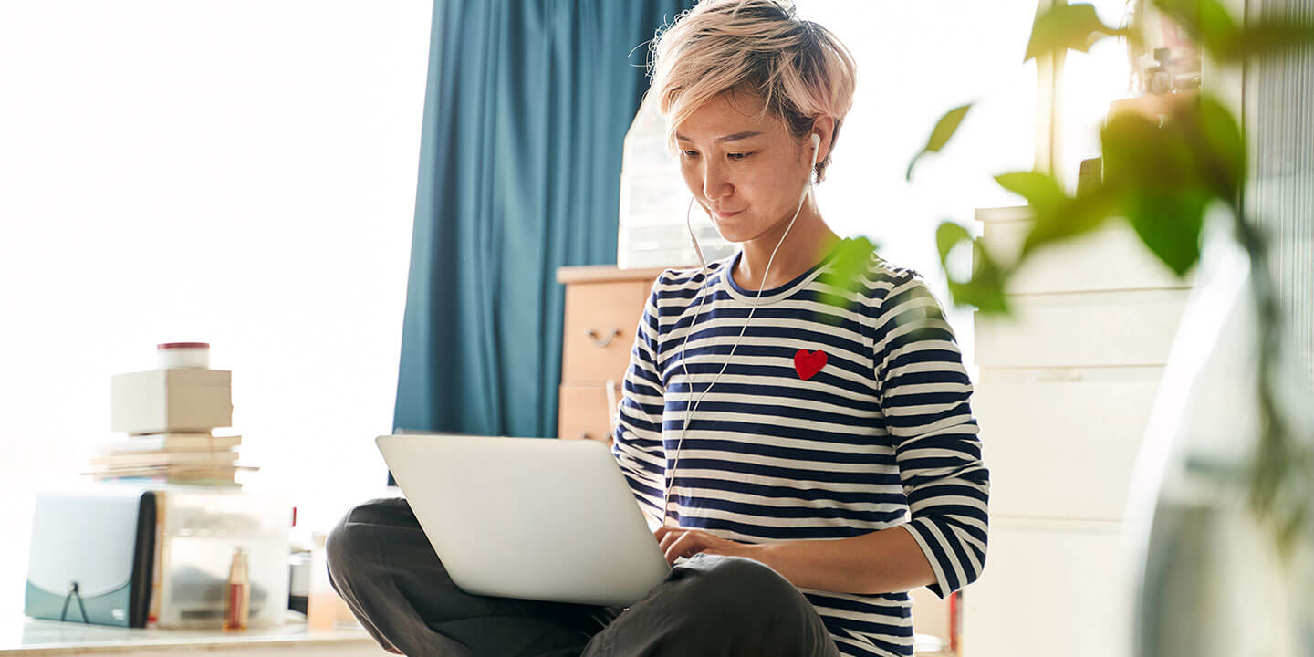 A woman sits cross-legged working on a laptop computer, earbuds in her ears.