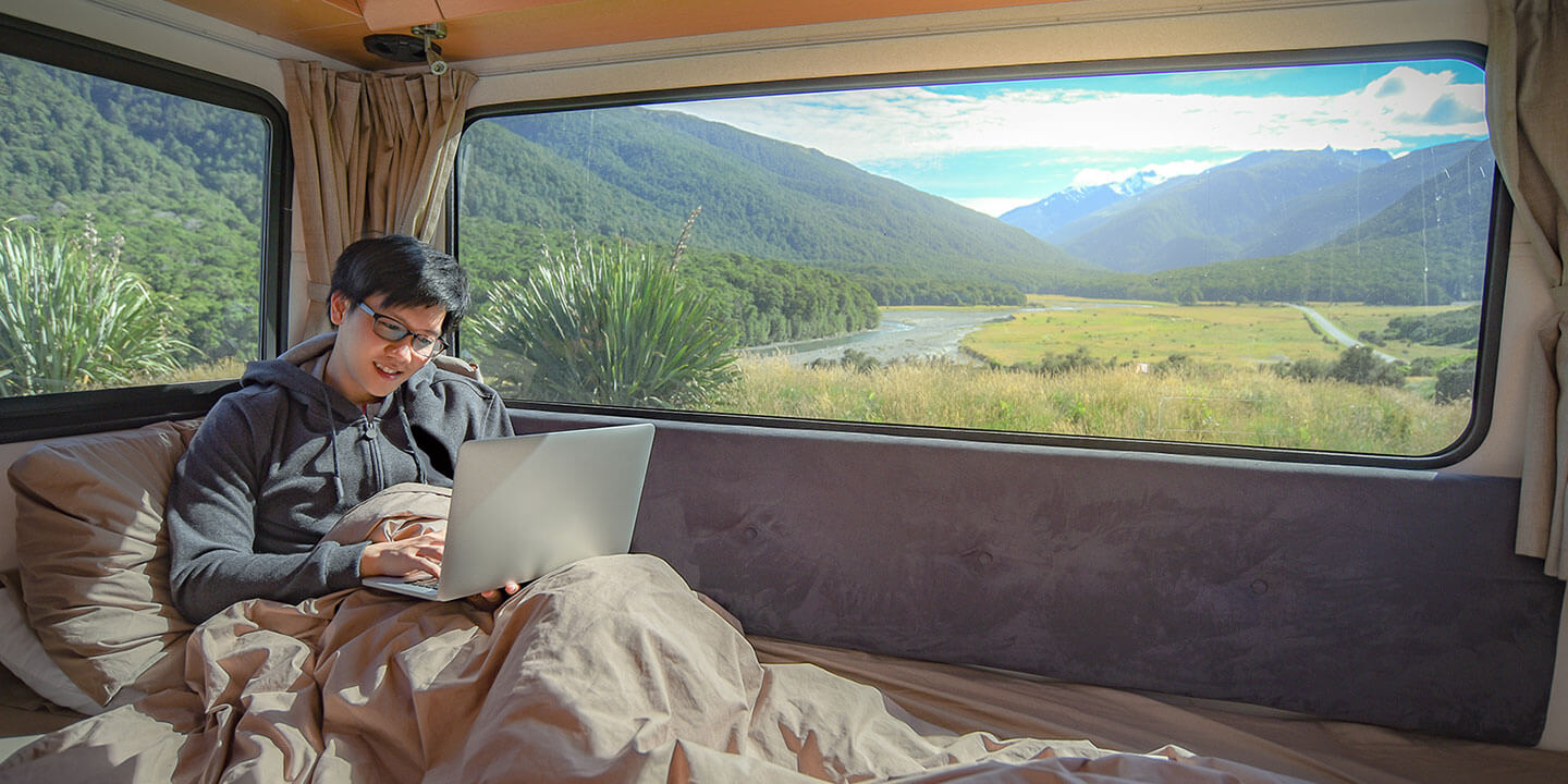 A man sits in a sleeping bag in an RV, mountains visible through the windows, while he works on his laptop computer