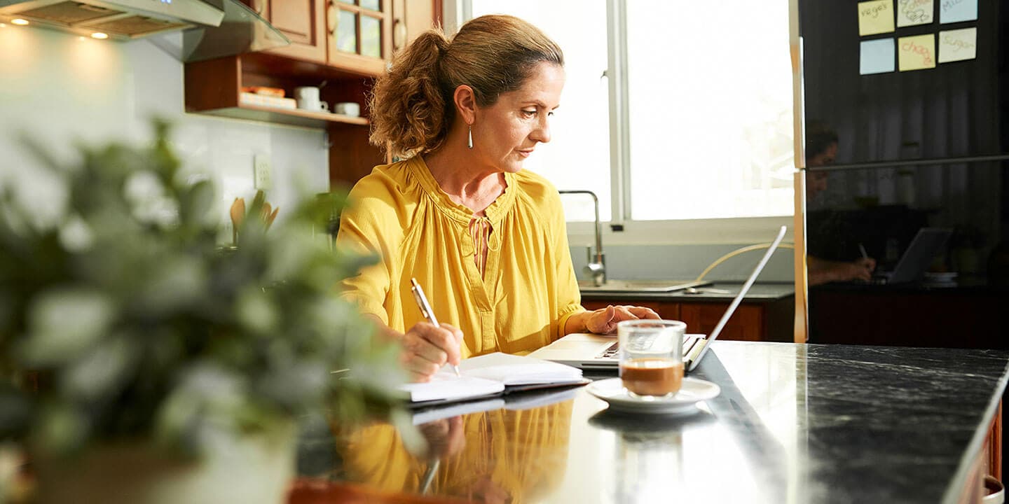 A woman sits at a laptop, writing in a notebook while looking at the screen.