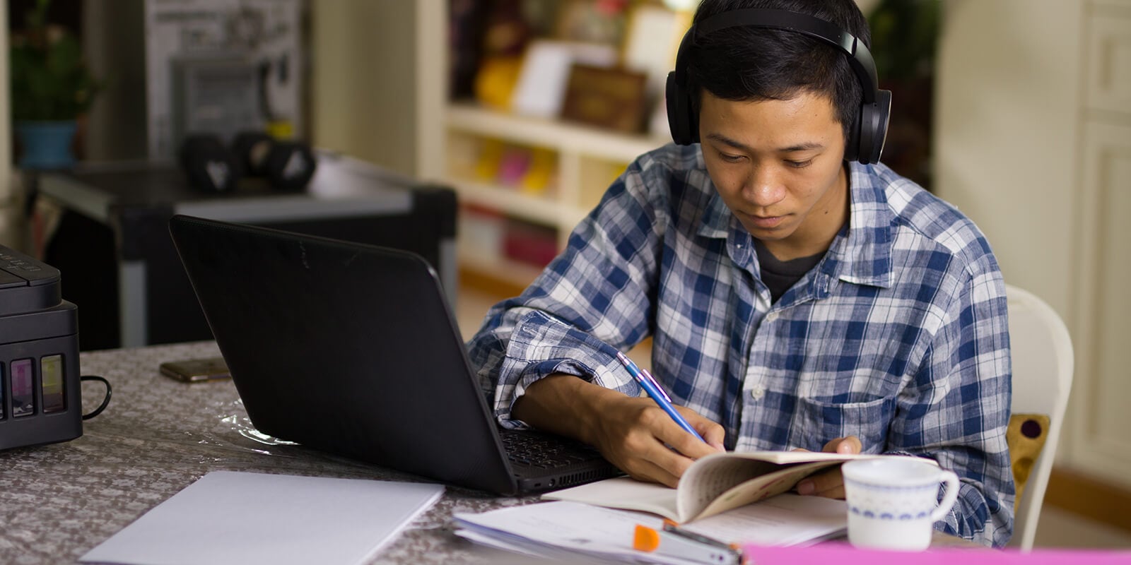 A man looking into a book, taking notes, with head phones on.