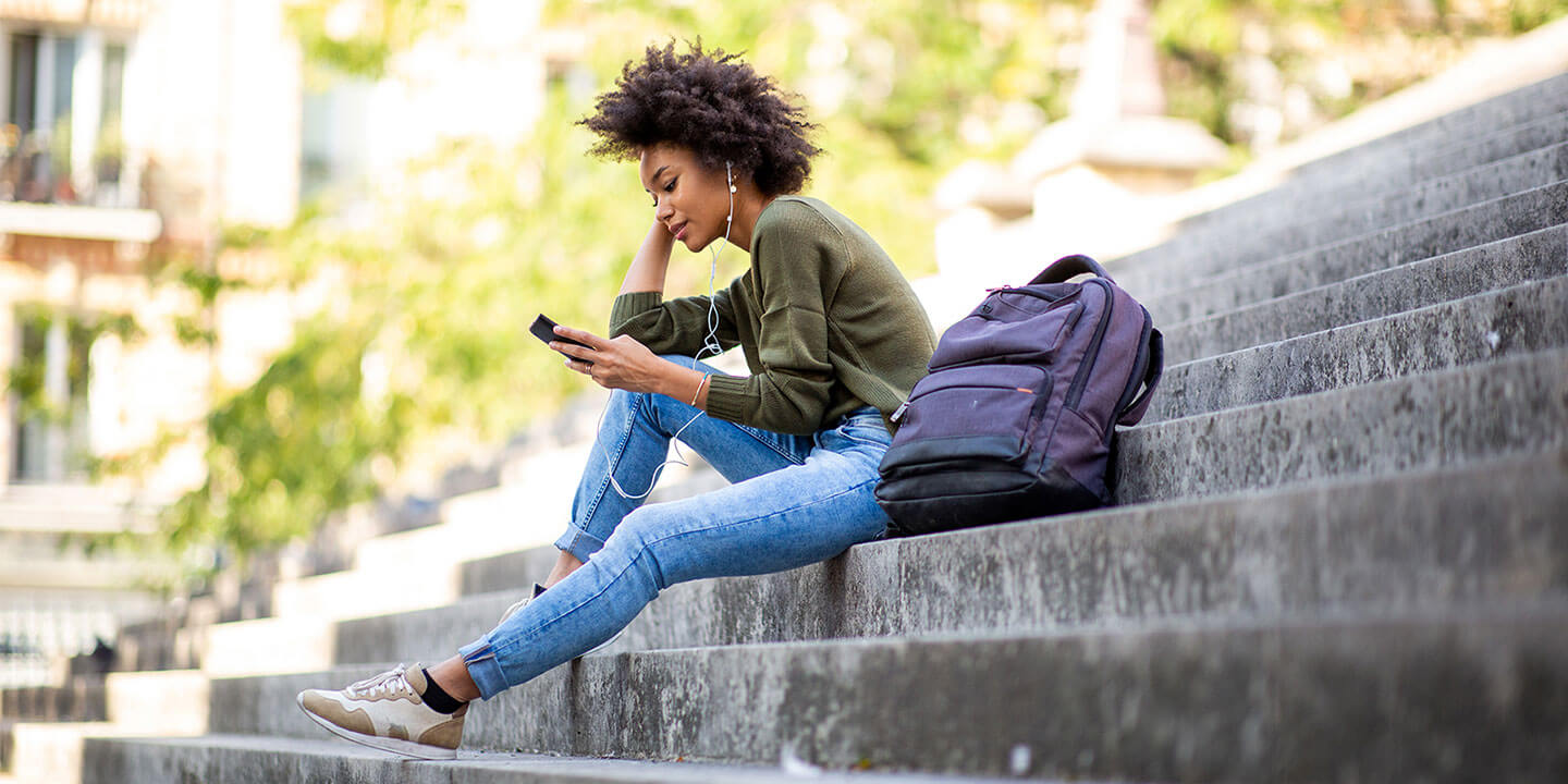 A woman sits on stone stairs, looking at her phone, with a backpack next to her.