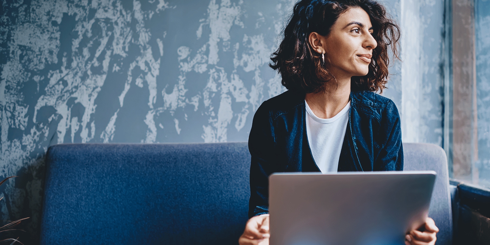 A woman looking out from a window with a happy face, holding laptop in her hands, sitting on a couch..