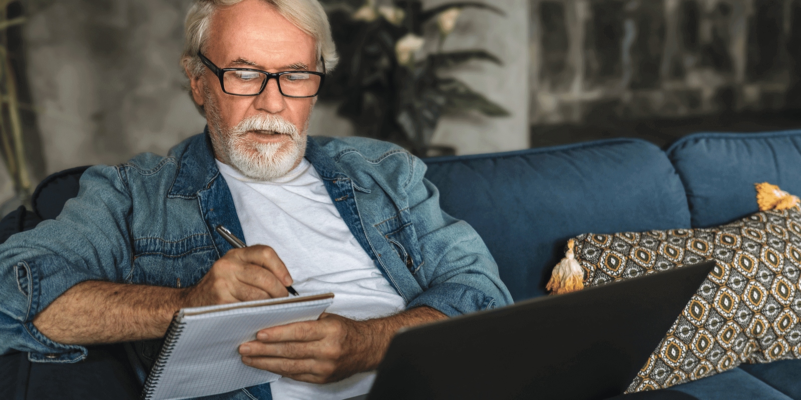 A man looking taking notes in a notebook, with a computer infront of him, sitting on a couch.