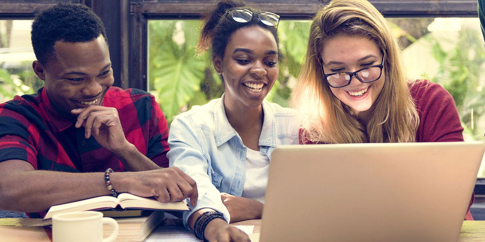 Two women and a man are sitting at a table that holds a laptop computer, books, and a mug. They are looking at the screen and smiling.