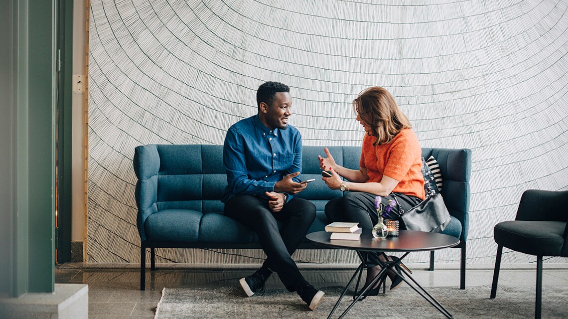 Two colleagues sitting on a sofa in a modern office lounge, discussing work while holding a smartphone and notebook at a small coffee table.