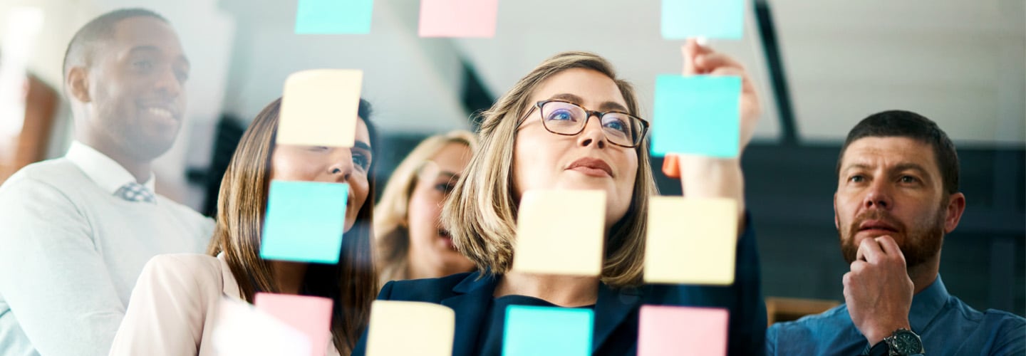 A business woman stood in front of a board of sticky notes, with others looking at the sticky notes.