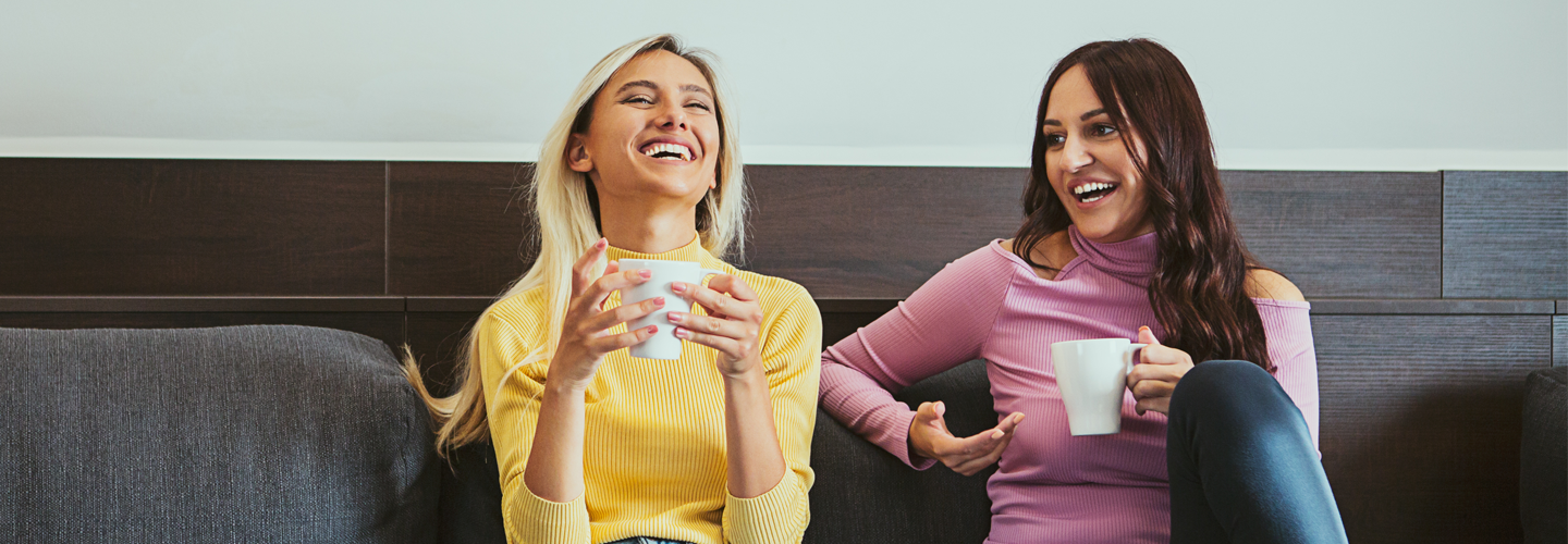 Two friends sat together on a sofa drinking hot drinks and smiling