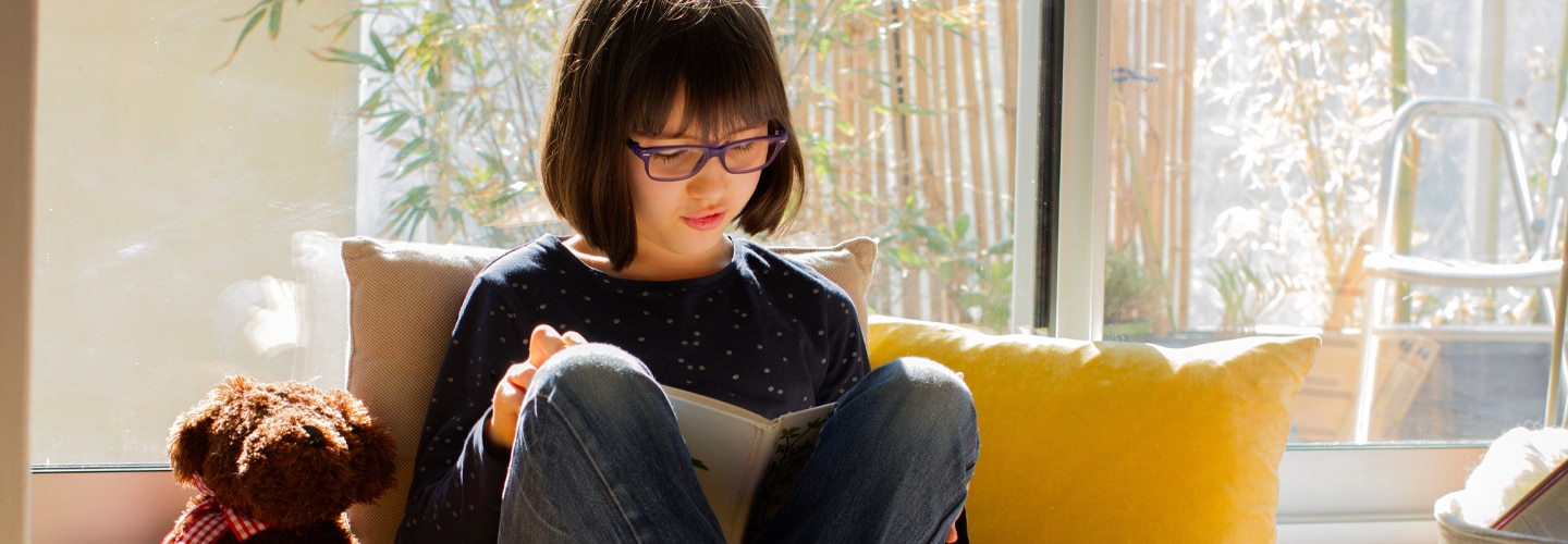 A girl sitting reading a book cross legged with a teddy bear next to her