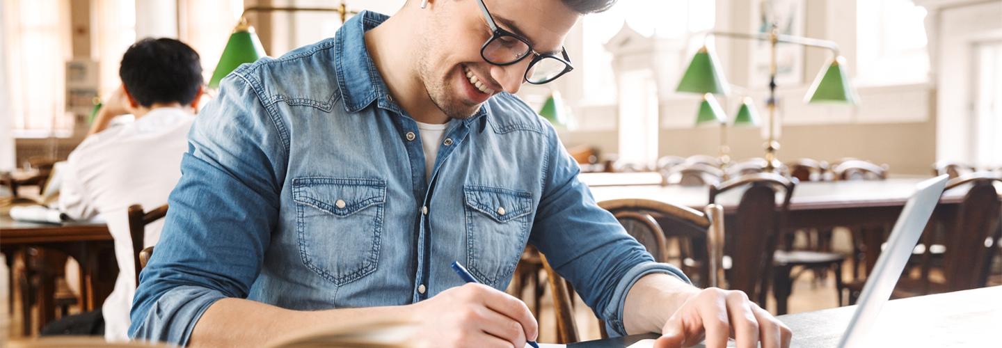 A man in a library with a open laptop and notepad studying and smiling