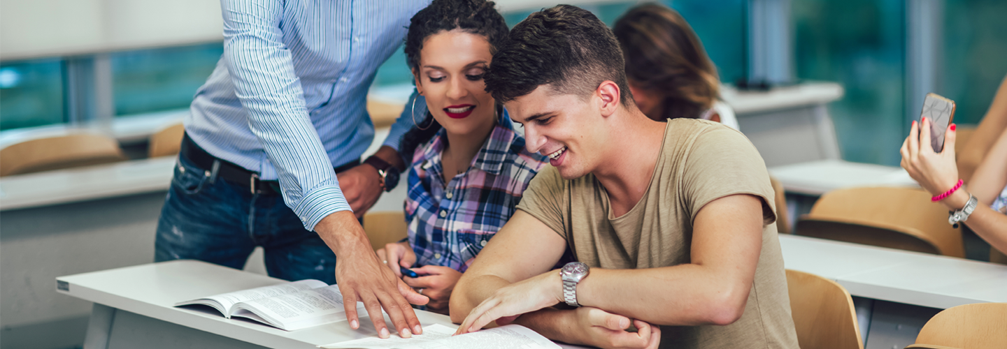 Students sat at a desk looking at a textbook together, with a teacher pointing to it