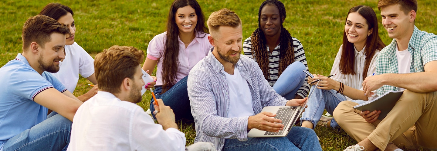 Students sat outside sat on grass, with a teacher in the middle on a laptop