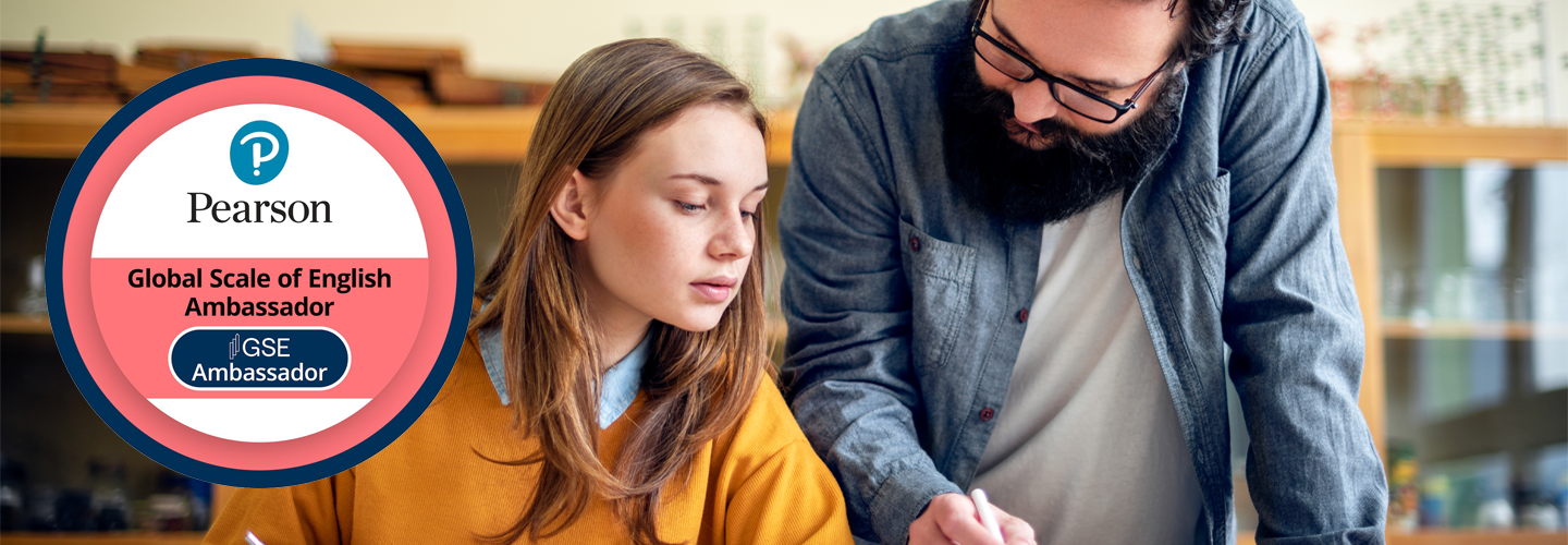 座った生徒の隣に立っているティーハーは、ペンを持っていて、テーブルの上の彼女の作品を身振りで示しています。左にはピンクの GSE アンバサダーのロゴ。