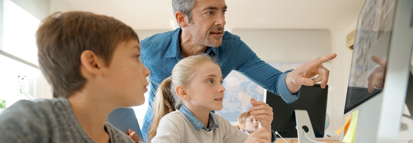 一人の教師が二人の幼い生徒のそばに立って、パソコンのモニターを見つめていた