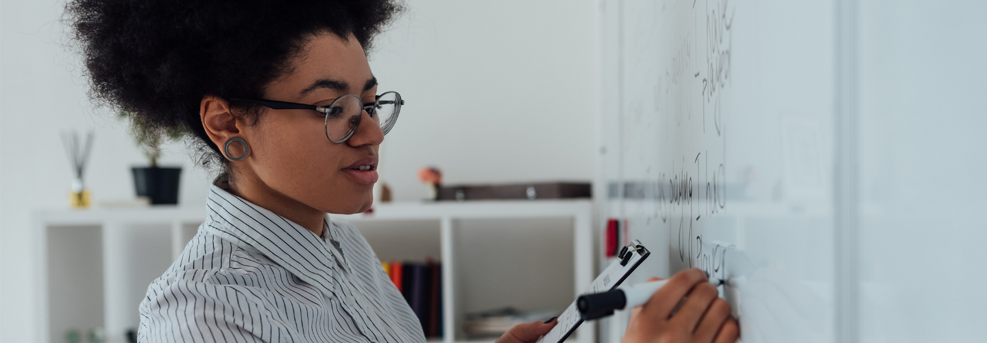 A person in a striped shirt writes with a marker on a whiteboard, holding a clip board
