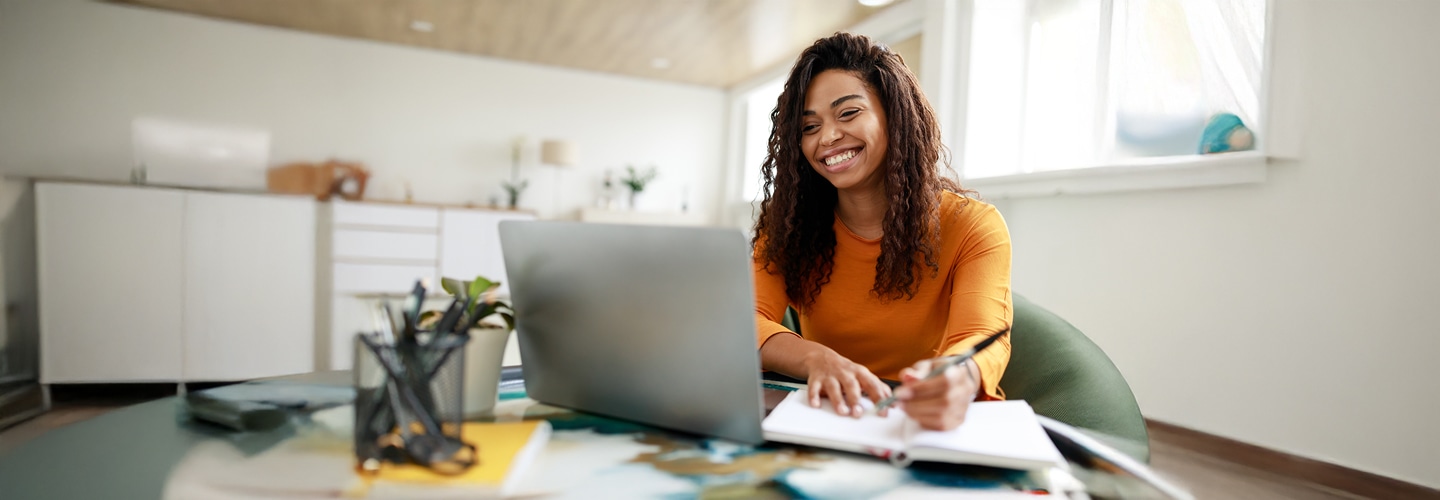woman writing in notepad while looking at laptop computer and smiling
